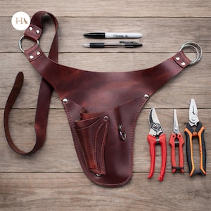Op de afbeelding: Een bruine leren tuingereedschapsriem met een schouderriem en twee metalen ringen. De riem heeft meerdere zakken en wordt vergezeld door een zwarte stift, een pen en drie tuingereedschappen met rode en oranje handvatten.