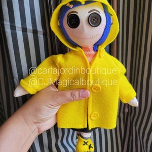 May include: A handmade doll wearing a yellow raincoat with a blue hood. The doll has large black button eyes and a stitched smile. The doll is holding a small yellow umbrella. The doll is standing on a striped background. @carlajordinboutique @CJMagicalboutique