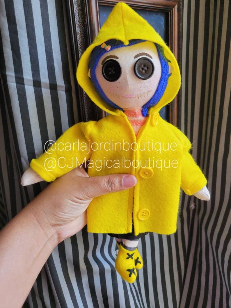 May include: A handmade doll wearing a yellow raincoat with a blue hood. The doll has large black button eyes and a stitched smile. The doll is holding a small yellow umbrella. The doll is standing on a striped background. @carlajordinboutique @CJMagicalboutique