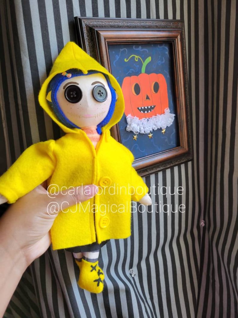 May include: A handmade doll wearing a yellow raincoat and black shoes. The doll has blue hair, a stitched-on smile, and large button eyes. The doll is standing in front of a framed picture of a jack-o'-lantern with a white lace collar. The picture has a blue background with gold stars. The text '@carlajordanboutique @CJMagicalBoutique' is visible in the image.