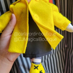 May include: A yellow felt doll wearing a yellow raincoat with black buttons and black shoes. The doll has a black skirt and white and black striped socks. The doll is holding a blue felt hat. The doll is standing on a black and white striped surface. @carlajordboutique @CJMagicalBoutique