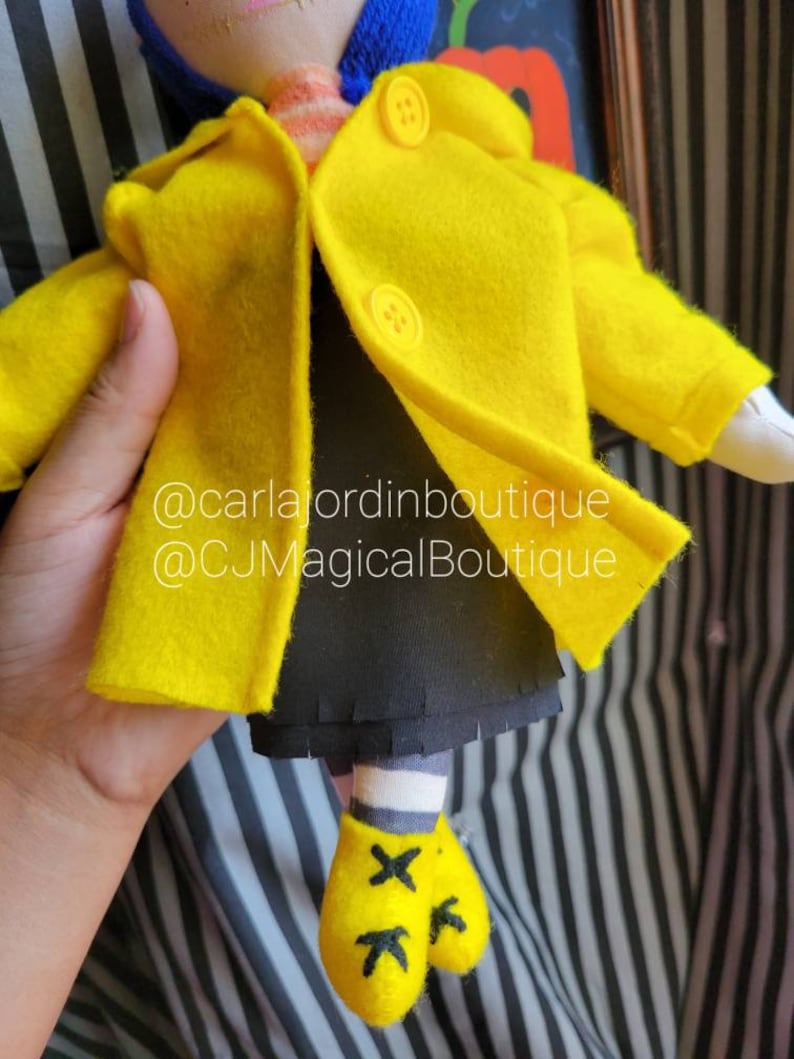 May include: A yellow felt doll wearing a yellow raincoat with black buttons and black shoes. The doll has a black skirt and white and black striped socks. The doll is holding a blue felt hat. The doll is standing on a black and white striped surface. @carlajordboutique @CJMagicalBoutique