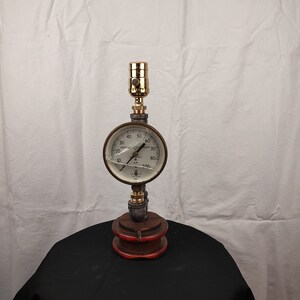 May include: A steampunk-style table lamp made from repurposed industrial parts. The lamp features a brass light fixture, a metal gauge with a white face and black markings, and a wooden base with a red accent.