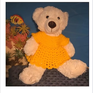 May include: A white teddy bear wearing a yellow crochet dress. The dress has a scalloped edge. The text at the top of the image reads "Teddy Bear Crochet Dress Pattern, 14" to 18" Size Bear."