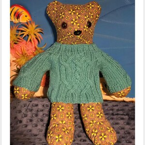 May include: A handmade teddy bear wearing a teal cable-knit sweater. The bear's head and paws are patterned with a brown floral design. The text "Whimsy Cable Bear Sweater Pattern" is at the top.