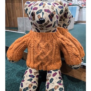 May include: A handmade teddy bear wearing an orange cable-knit sweater. The bear's head and feet are patterned with colorful mittens and other shapes. The text "Sassy Cable Bear Sweater Pattern" is at the top.