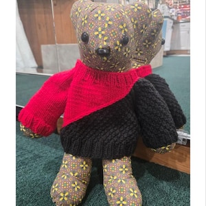 Könnte beinhalten: Eine handgefertigte Bärenpuppe mit gemustertem Kopf und Füßen, die einen diagonalen rot-schwarzen Pullover trägt. Der Text "2 Color Diagonal Team Bear Sweater" befindet sich oben im Bild. Der Bär hat schwarze Knopfaugen und eine schwarze Nase.