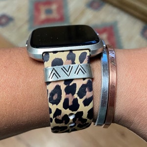 May include: A smartwatch with a leopard print band and a silver metal band with the letters "AVA". Two silver and copper bracelets are also visible.