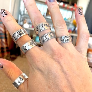 May include: A hand wearing five silver rings with different designs. The rings feature the words "Long Live Cowboys", "Yee Haw", "1st Buck Off", and a cactus design. One ring has a geometric pattern.