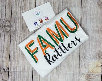 May include: White t-shirt with the text "FAMU Rattlers" in green, orange, and black lettering. The letters are outlined in orange and green.