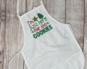 Puede incluir: Delantal blanco con un diseño bordado en verde y rojo que dice "Solo estoy aquí por las galletas" con una estrella y un árbol de Navidad.