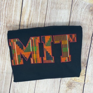 Könnte beinhalten: Schwarzes Sweatshirt mit dem Wort "MIT" in großen, bunten Buchstaben. Die Buchstaben bestehen aus einem gemusterten Stoff in den Farben Orange, Grün und Gelb.