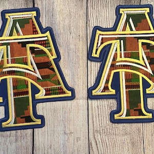 May include: Two embroidered patches featuring the letters "AT" in a stylized design. The letters are filled with a colorful African print fabric, outlined in gold and white, and set against a blue background. The patches are on a light wood grain surface.