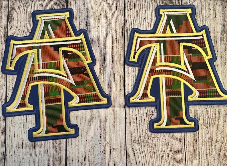May include: Two embroidered patches featuring the letters "AT" in a stylized design. The letters are filled with a colorful African print fabric, outlined in gold and white, and set against a blue background. The patches are on a light wood grain surface.