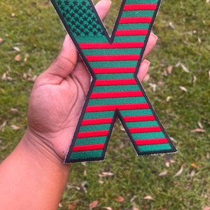 May include: A red and green embroidered iron-on patch shaped like the letter X. The patch is designed to look like an American flag, but with red and green stripes instead of red, white, and blue.