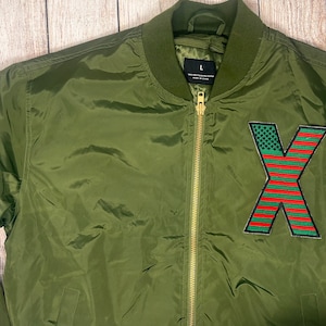 May include: A green bomber jacket with a large red, white, and blue "X" patch on the front. The zipper is gold.