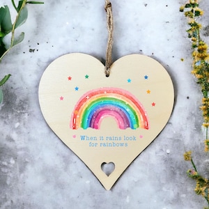 A wooden hanging gift heart with aheart cut out at the bottom. The design printed onto the sign is a rainbow with rainbow coloured stars and the quote When it rains look for rainbows.