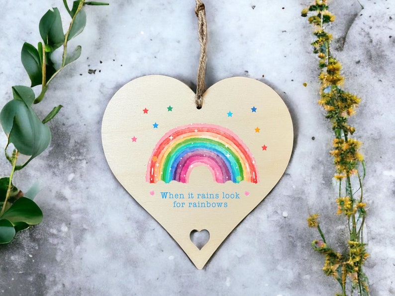 A wooden hanging gift heart with aheart cut out at the bottom. The design printed onto the sign is a rainbow with rainbow coloured stars and the quote When it rains look for rainbows.