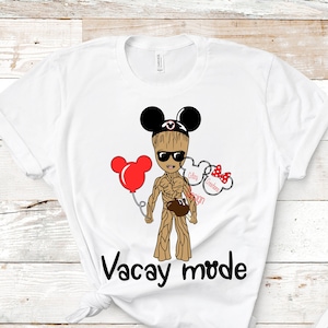 May include: White t-shirt with a cartoon image of a brown character wearing Mickey Mouse ears and sunglasses. The character is holding a red balloon and a brown object. The text "Vacay mode" is printed below the character.