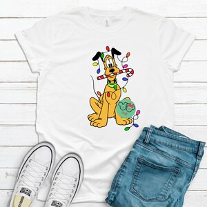 Peut inclure: Un t-shirt blanc avec une image de dessin animé de Pluto le chien portant une canne en sucre et entouré de lumières de Noël.