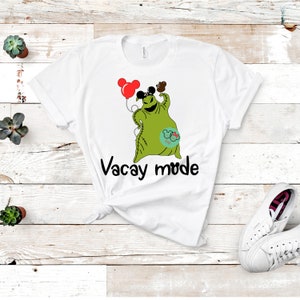 Puede incluir: Camiseta blanca con un personaje de dibujos animados verde que sostiene un globo y lleva un sombrero de orejas de Mickey Mouse. El texto "Vacay mode" está impreso debajo del personaje.