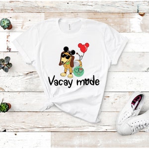Può includere: Maglietta bianca con un cane di cartone animato che indossa occhiali da sole e orecchie di Topolino. Il cane tiene un palloncino rosso con orecchie di Topolino. Il testo "Vacay mode" è stampato sotto il cane.