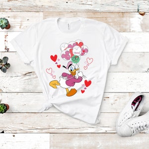 Puede incluir: Camiseta blanca con un pato de dibujos animados rosa y blanco que sostiene globos en forma de corazón rosa y rojo. Los globos tienen la palabra "XOXO" en ellos. El pato lleva un vestido rosa y un sombrero blanco. Hay pequeños corazones rosas esparcidos alrededor del pato.