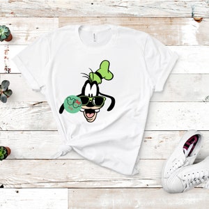 May include: White t-shirt featuring a cartoon image of Goofy wearing sunglasses and a green hat. The design includes a small, round graphic with text. The shirt is styled with a knot at the waist. A pair of white sneakers is visible in the bottom right corner.