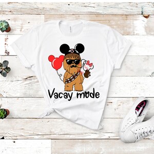 May include: White t-shirt with a cartoon image of Chewbacca wearing Mickey Mouse ears and sunglasses. The image also includes a red balloon and a Mickey Mouse head with a bow. The text "Vacay mode" is printed below the image.