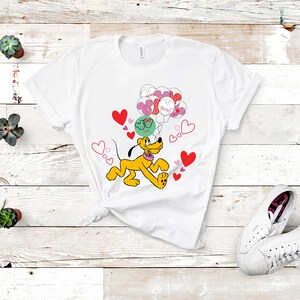 Puede incluir: Camiseta blanca con un perro amarillo de dibujos animados que sostiene globos en forma de corazón rosa y rojo. Los globos tienen el texto "You're" y "Great!" El perro está corriendo y hay corazones esparcidos a su alrededor.