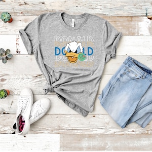 May include: A gray t-shirt with a graphic of Donald Duck's face and the word "DONALD" repeated in a stacked pattern. The shirt is laid out on a wooden surface with a pair of white sneakers and blue jeans.
