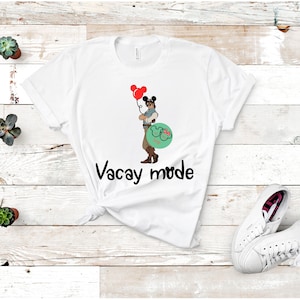 May include: White t-shirt with a cartoon image of a man wearing a cowboy hat and holding red balloons. The man is wearing a blue shirt and brown pants. The text "Vacay Mode" is printed below the image.