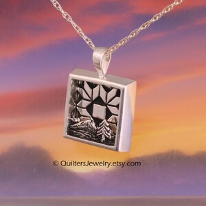 May include: A silver pendant necklace with a square frame design. The pendant features a geometric star pattern and a landscape scene with mountains and trees. The necklace is suspended from a delicate silver chain. The text "QuiltersJewelry.etsy.com" is visible.