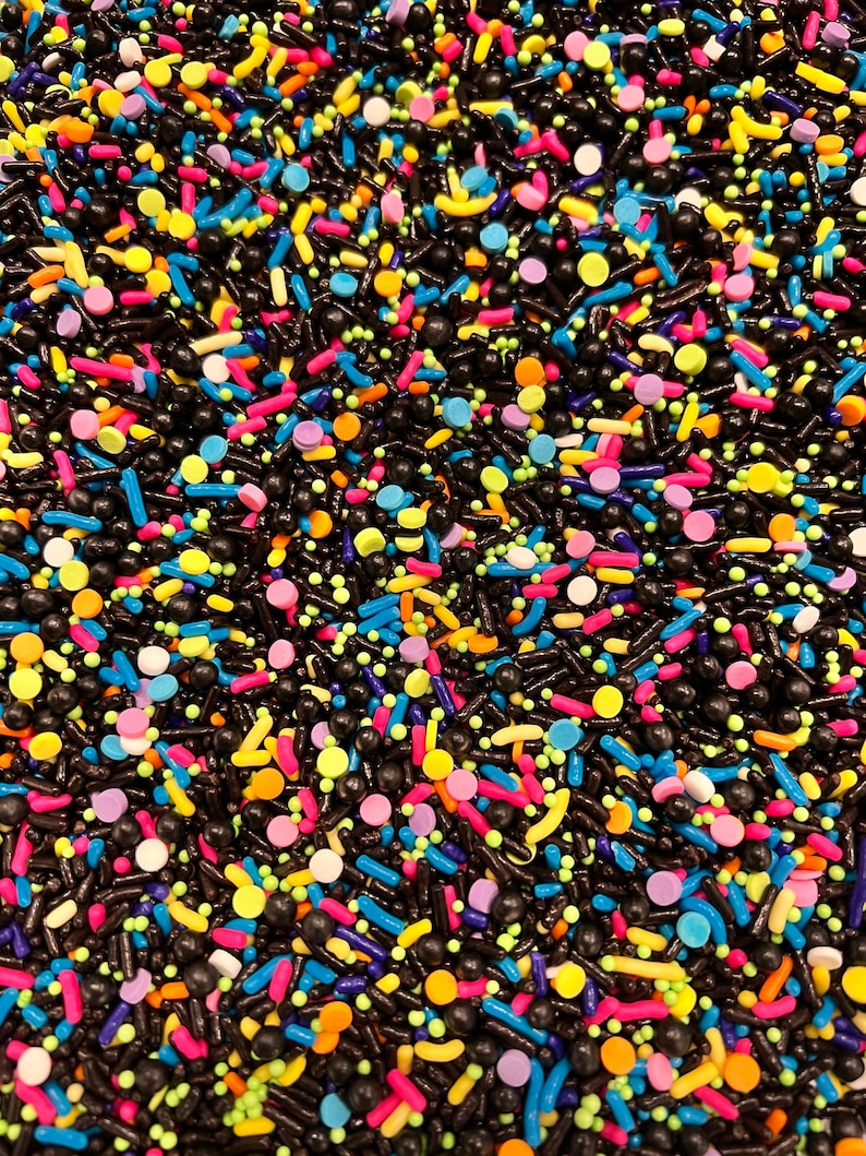 May include: A close-up of a variety of colourful sprinkles, including black, yellow, blue, green, pink, orange, and white. The sprinkles are a mix of shapes, including long, thin sticks, small balls, and flat discs.