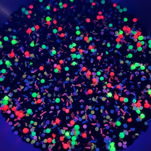 May include: A close-up of a mixture of black, red, green, and blue sprinkles. The sprinkles are in a bowl and are glowing under a black light.