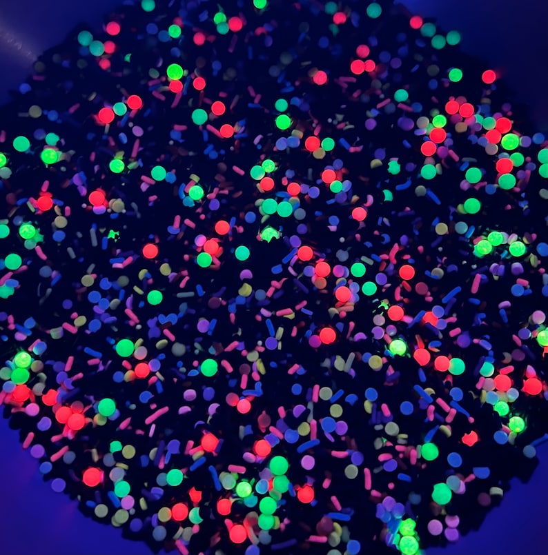 May include: A close-up of a mixture of black, red, green, and blue sprinkles. The sprinkles are in a bowl and are glowing under a black light.