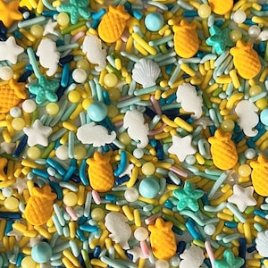 May include: A mix of colorful sprinkles in blue, yellow, and green. The sprinkles are shaped like pineapples, seashells, starfish, and seahorses.