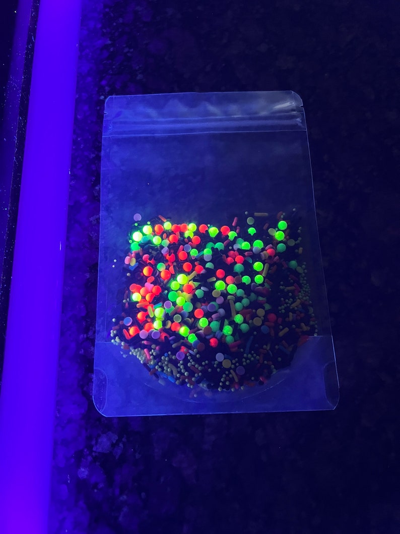 May include: A clear plastic bag filled with colourful sprinkles. The sprinkles are a mix of red, green, yellow, and black. The sprinkles are in a variety of shapes and sizes.