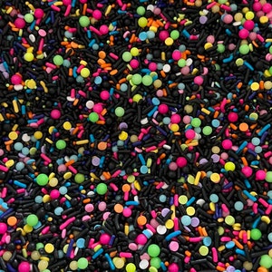 May include: A close-up image of colourful sprinkles, including black, pink, green, blue, orange, and yellow. The sprinkles are a mix of round and long shapes.