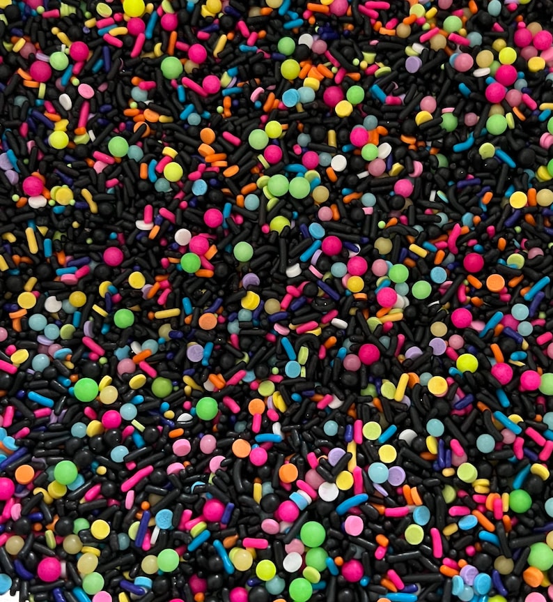 May include: A close-up image of colourful sprinkles, including black, pink, green, blue, orange, and yellow. The sprinkles are a mix of round and long shapes.