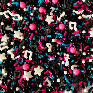Pode incluir: Uma mistura de confeitos pretos, brancos, rosa e azuis em forma de estrelas, notas musicais e pequenos cilindros.