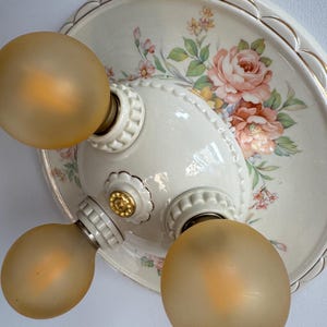 May include: A round, cream-colored ceramic ceiling light fixture featuring a scalloped edge with gold trim. The surface is decorated with a floral pattern of pink roses and green leaves. It includes three sockets with frosted, amber-toned bulbs.