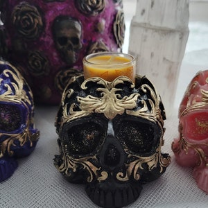 May include: Three decorative skull candle holders with gold accents. Each skull is a different color: purple, black, and pink. Each skull has a small glass votive candle holder on top.