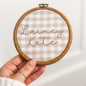 May include: A brown wooden embroidery hoop with a white and beige gingham fabric inside. The fabric has the names "Lainey Kate" embroidered in brown thread.