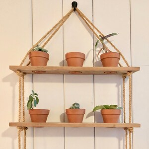 Może przedstawiać: Trójpoziomowa drewniana półka z linowymi wieszakami, na której stoją różne rośliny doniczkowe. Półki są wykonane z naturalnego drewna, a lina jest jasnobrązowa. Rośliny to różne sukulenty i inne małe rośliny.