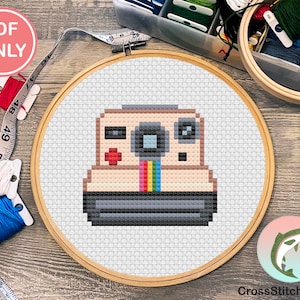 Op de afbeelding: Een kruissteekpatroon van een vintage camera met een regenboogfilmstrip. Het patroon is in een houten borduurring op een houten oppervlak. De tekst "CrossStitchSalmon" is zichtbaar in de rechterbenedenhoek van de afbeelding.