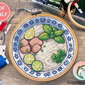 May include: A cross-stitch embroidery hoop featuring a detailed design of a bowl of pho. The design includes noodles, meat, lime wedges, and herbs, all within a blue and white patterned border. The text "PDF ONLY" is visible.