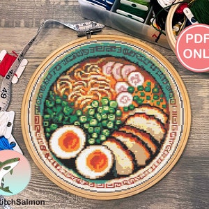 May include: A cross stitch pattern of a bowl of ramen noodles. The pattern is in a wooden embroidery hoop and is ready to be stitched. The pattern includes noodles, broth, pork, eggs, green onions, and other toppings.