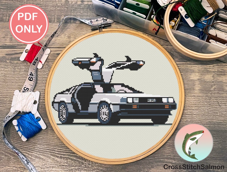 May include: A cross stitch pattern of the DeLorean time machine from the Back to the Future movie. The car is white with a black front and a gray interior. The time machine is flying with its doors open. The pattern is in a round wooden embroidery hoop.