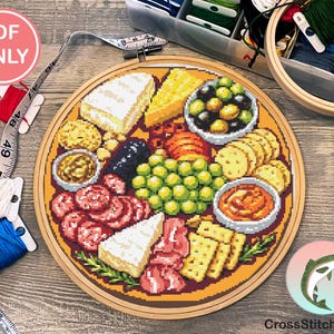 May include: A cross-stitch pattern of a charcuterie board, featuring cheese, olives, crackers, and meats. The design is set in a wooden embroidery hoop. The image includes spools of thread, a measuring tape, and the text "PDF ONLY".